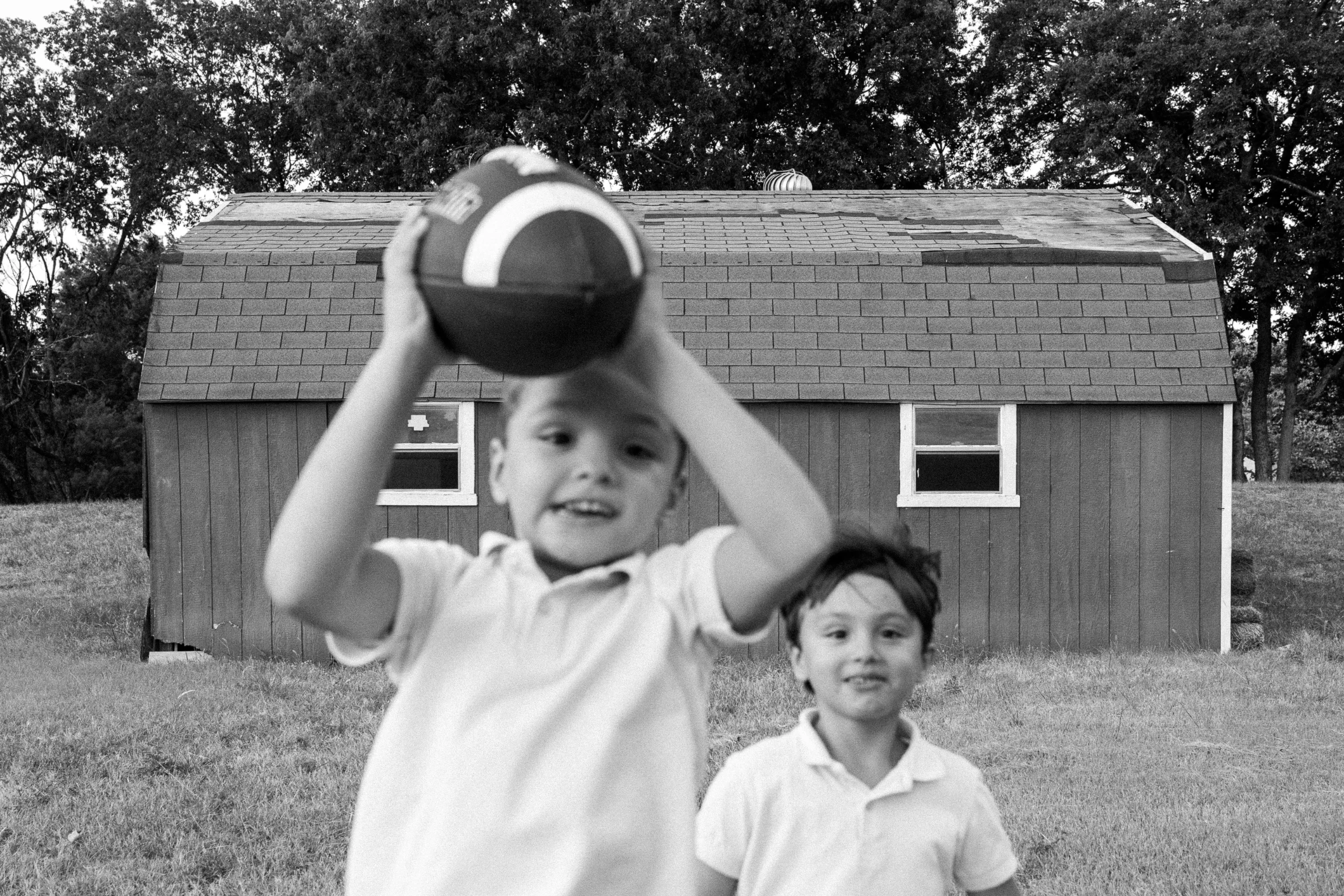 Black and white photo of two boys in polo shirts playing with a football outside a barn during a family session.