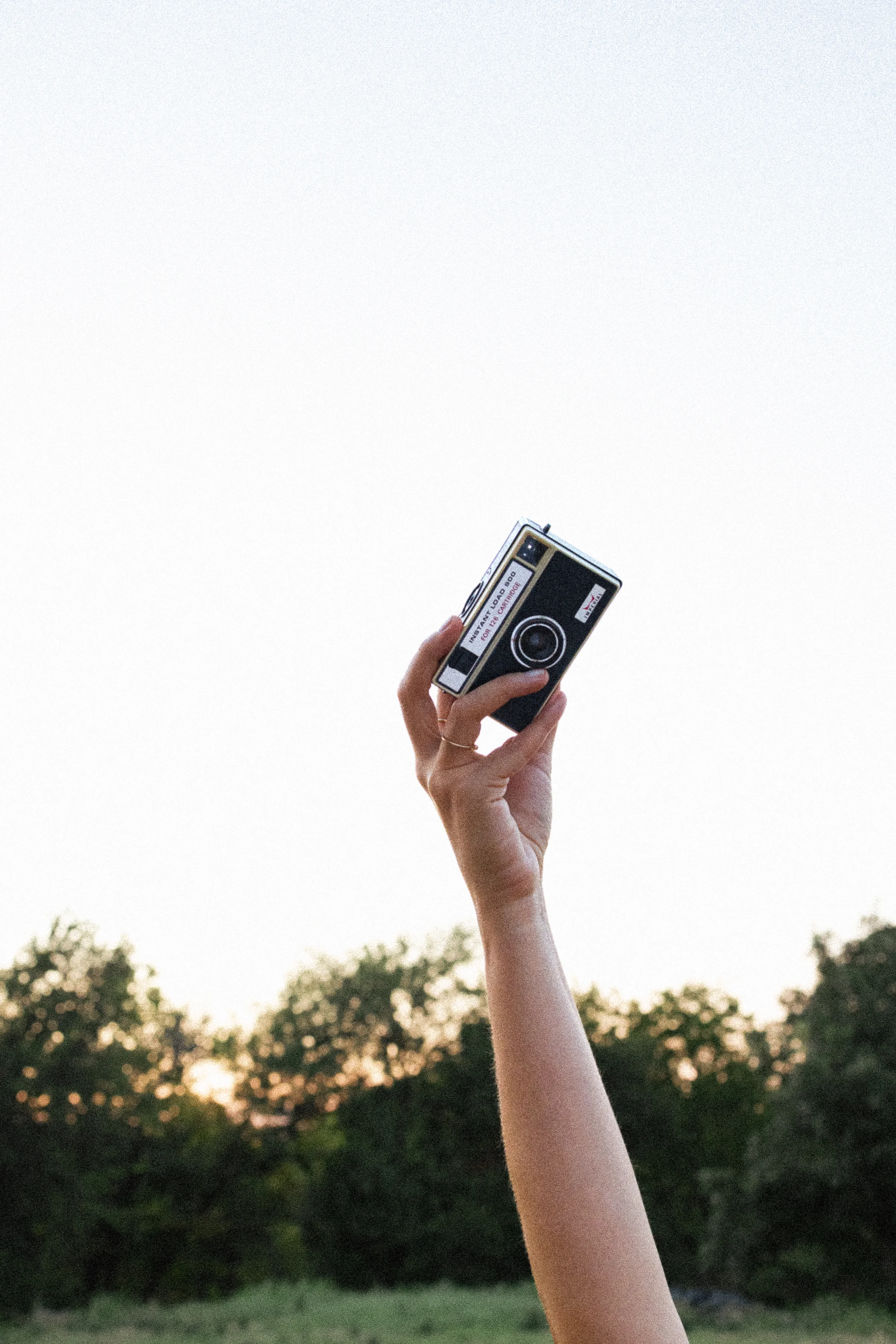 Hand raised holding a vintage film camera against a pale sky above a treeline at golden hour.