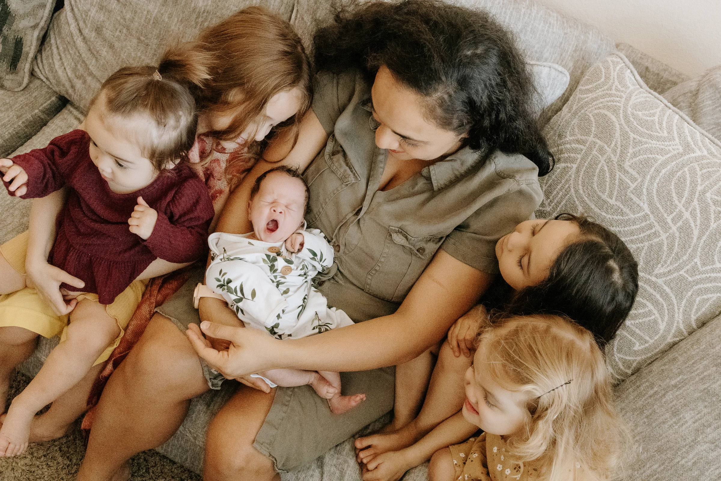Mother in olive dress holding her yawning newborn on a couch surrounded by four young children, in home family session.