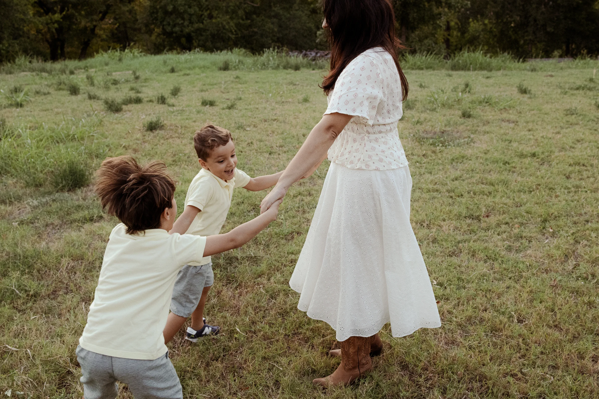 Mother in a white dress holding hands and playing in a ring with two boys in yellow shirts in a grassy field.