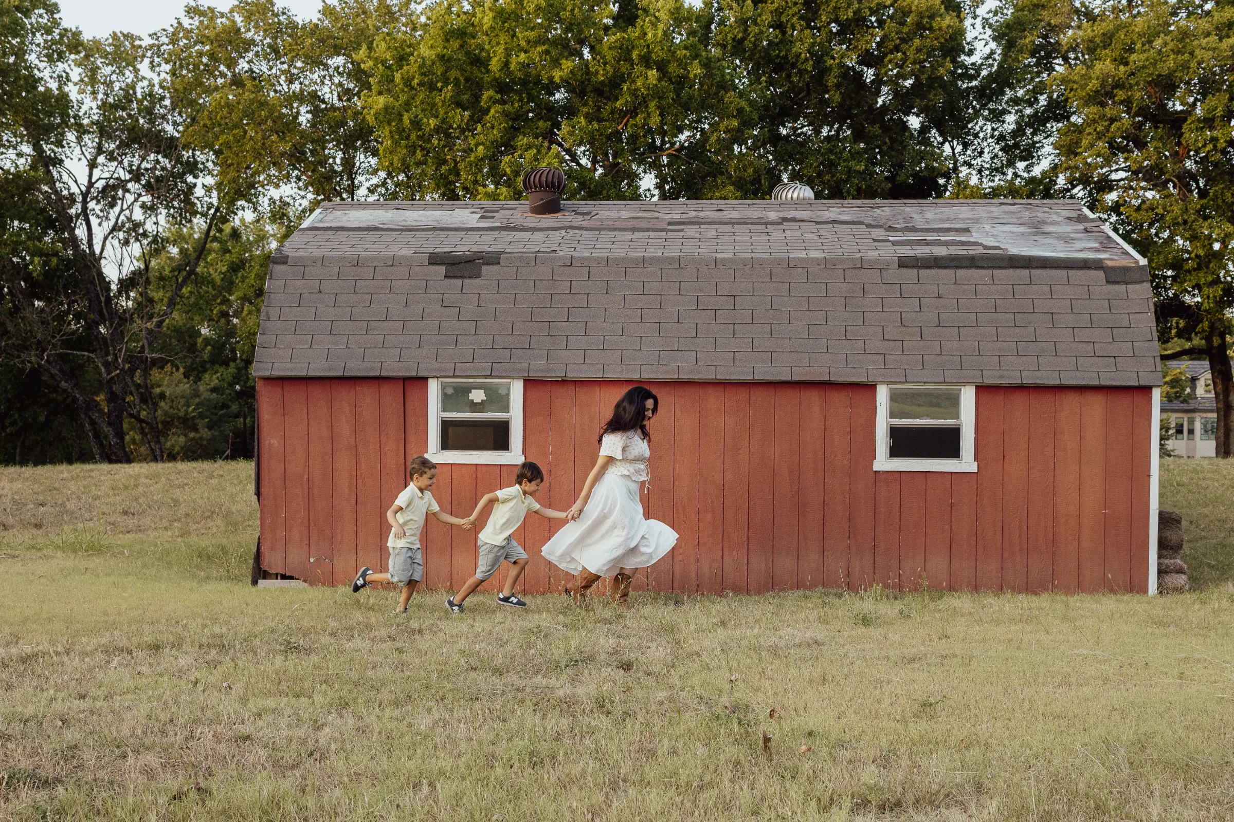 Mother in a white dress holding hands with two boys walking past a red barn in a grassy field during a family session.