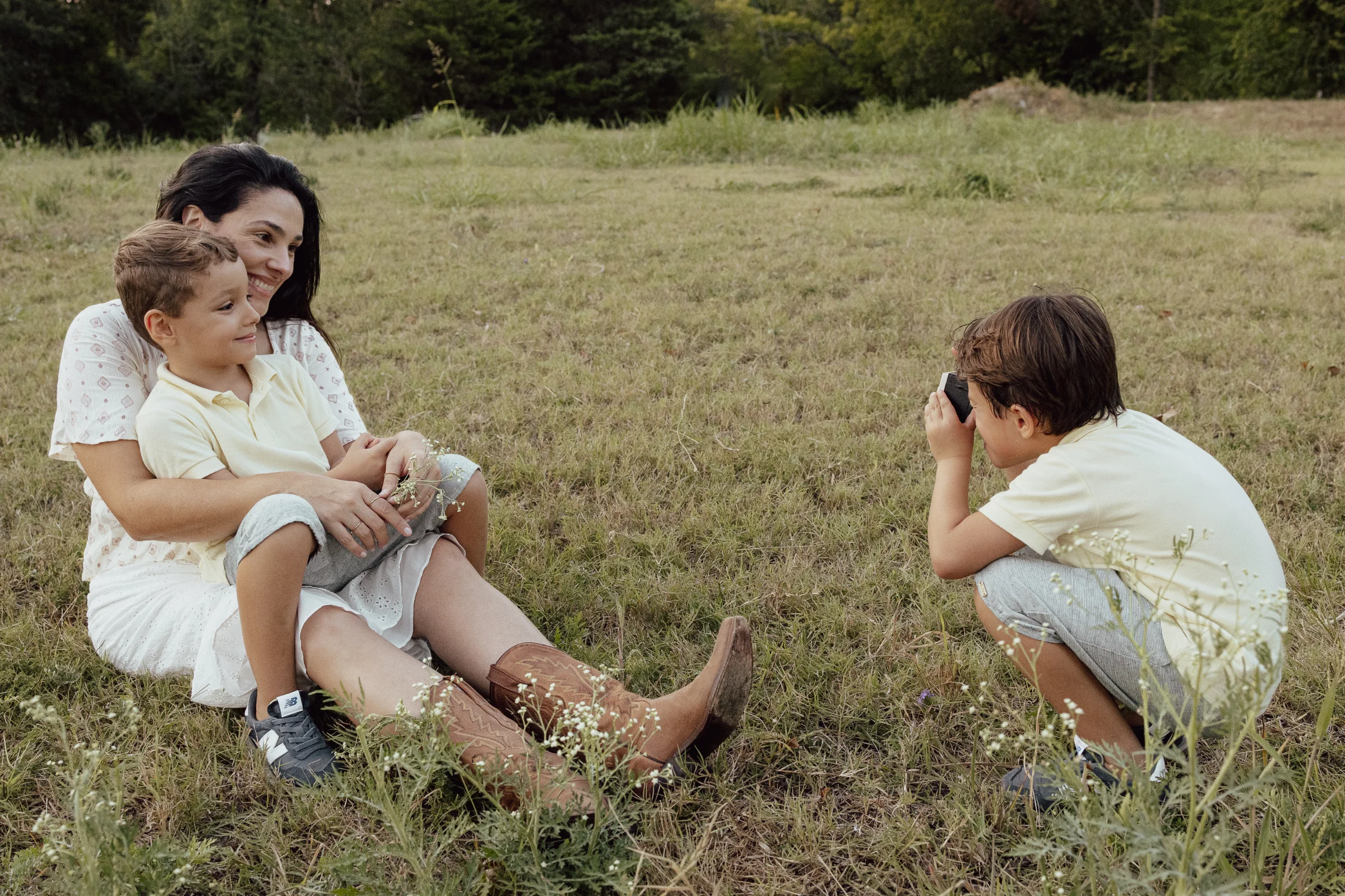 Mother in white dress holding a young boy in a grassy field as another boy photographs them with a camera.