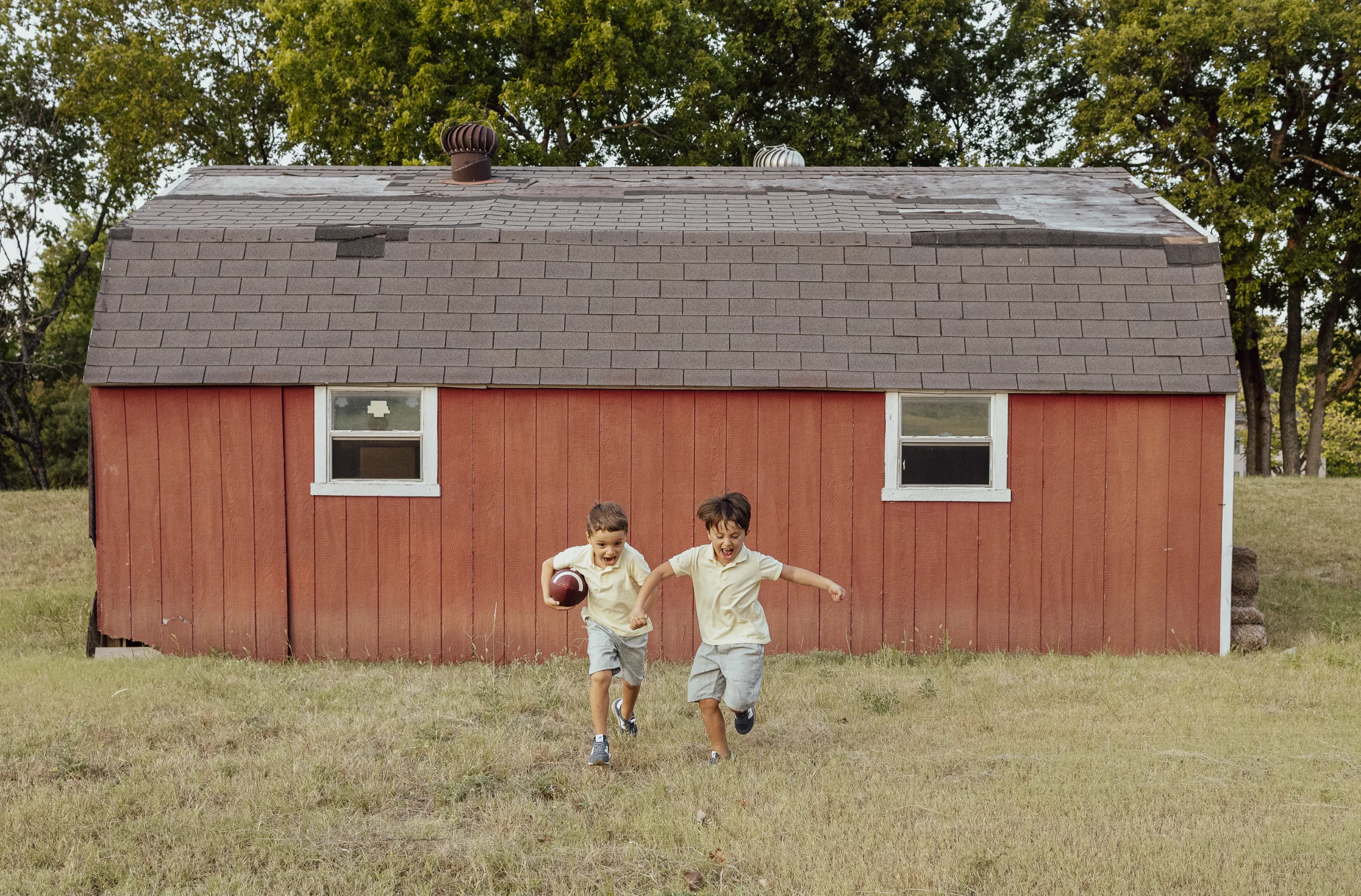 Two boys in yellow shirts run laughing with a football past a red barn in a grassy field, family session