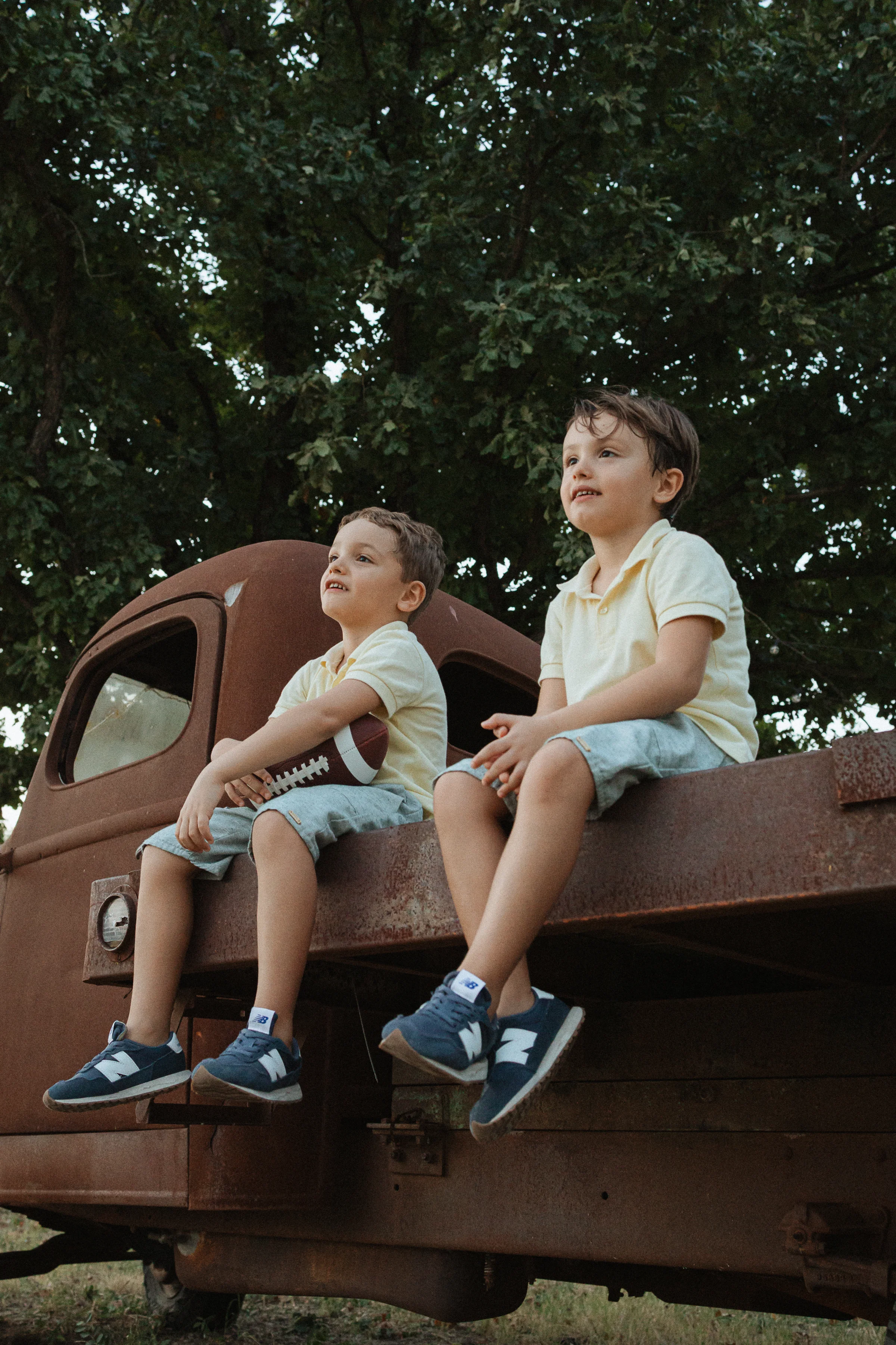 Two young boys in yellow polos and denim shorts sit on a rusty vintage truck holding a football outdoors.