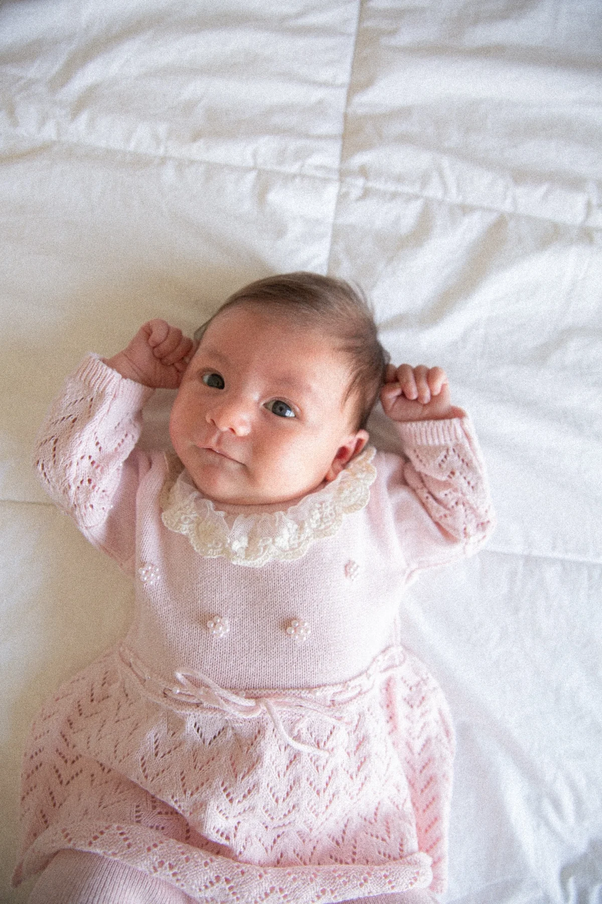 Newborn baby awake in a pink knit dress, resting on white bedding